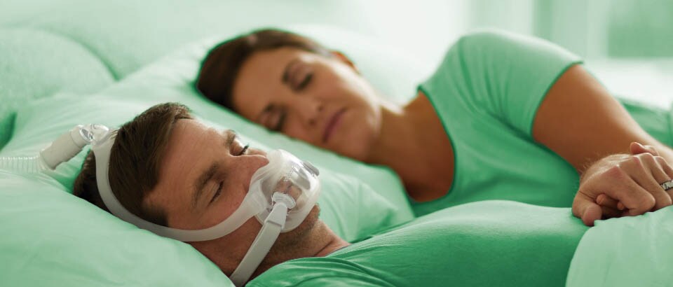 Hombre con camiseta verde durmiendo con una mascarilla, y mujer con camiseta verde durmiendo a su lado.