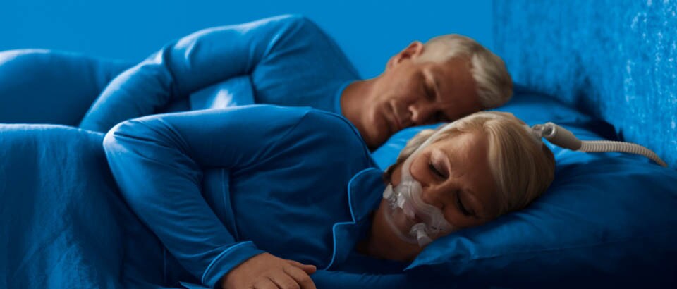 Una mujer vestida de azul durmiendo de lado con una mascarilla, y un hombre también vestido de azul durmiendo de lado.